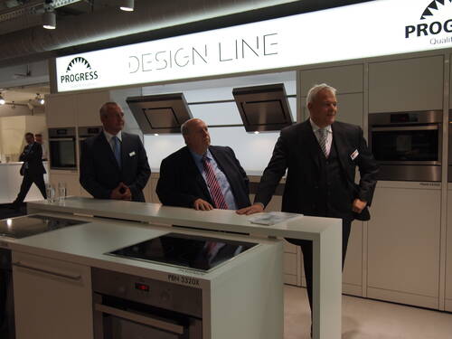 Drei Geschäftsleute in Anzügen präsentieren moderne Einbauküchen und Kochfelder an einem Messestand der Marke „Progress Design Line“.