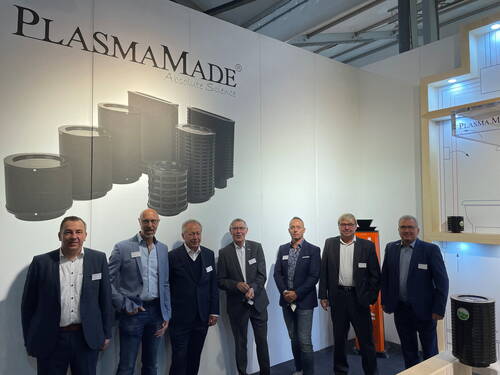 Sieben Geschäftsleute in Anzügen stehen nebeneinander vor einem Messestand von PlasmaMade mit großformatigen Abbildungen von Filtermodulen an der Wand.
