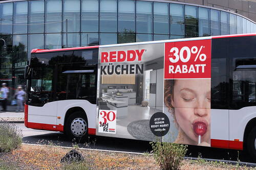 Werbebus mit großflächigem Reddy-Küchenplakat, das eine moderne Einbauküche, eine lachende Frau mit Lolli und den Hinweis auf bis zu 30 Prozent Rabatt zeigt.