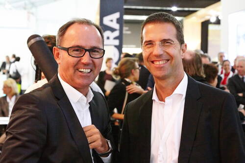 Zwei lächelnde Geschäftsmänner im Anzug unterhalten sich auf einer gut besuchten Messe oder Konferenz im Innenraum.