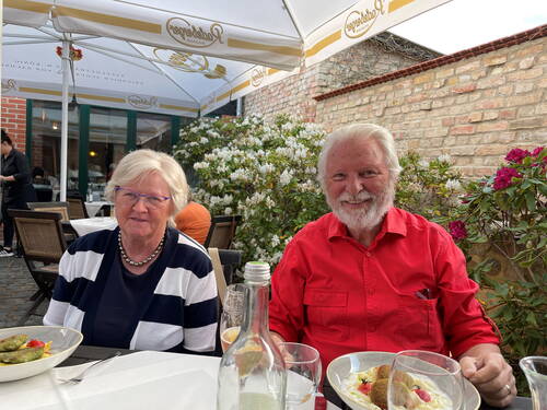 Älteres Paar sitzt lächelnd an einem Restauranttisch im Freien und genießt ihr Essen zwischen blühenden Sträuchern unter großen Sonnenschirmen.
