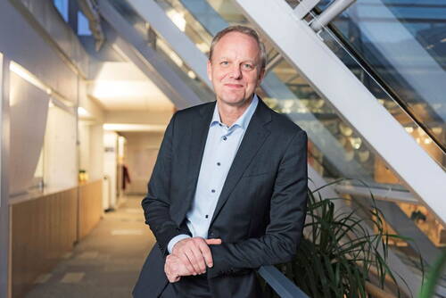 Mittelalterlicher Mann im Anzug lehnt sich lächelnd an ein Treppengeländer in einem modernen, hell beleuchteten Bürogebäude