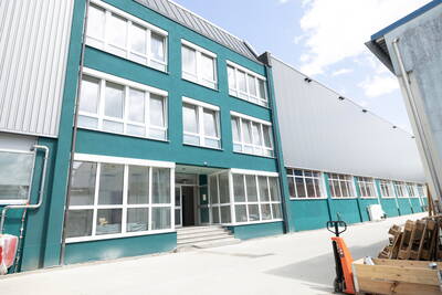 Modernes, türkisfarbenes Industrie- und Bürogebäude mit großen weißen Fenstern und seitlichem Lagerbereich mit Paletten und Hubwagen im Hof