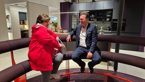 Zwei Menschen sitzen einander zugewandt auf einer runden Polsterbank in einer modernen Ausstellungsküche und unterhalten sich lächelnd.