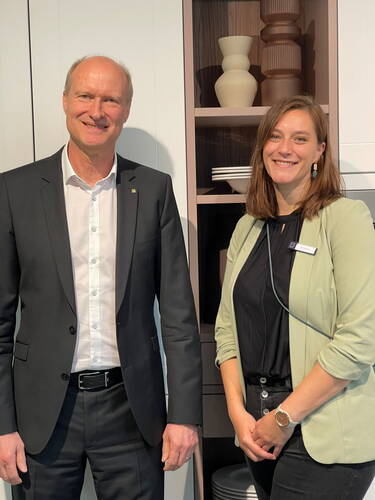 Zwei lächelnde Personen in Businesskleidung stehen nebeneinander vor einem Regal mit dekorativen Vasen und Tellern