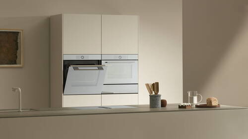 Minimalistische, beige Küche mit geöffnetem Einbau-Backofen, aufgeräumter Arbeitsplatte mit Kochfeld, Utensilienbehälter, Brotlaib und Glaskaraffe mit Wasser.