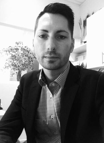 Junger Mann im Anzug sitzt in einem Büro und blickt ruhig in die Kamera, im Hintergrund ein großer Zimmerpflanze und Regale mit Unterlagen.