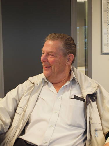 Älterer Mann mit zurückgekämmten Haaren sitzt lächelnd in einem Büro und trägt ein offenes, helles Jackett über einem weißen Hemd.