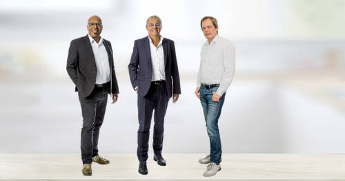 Drei Männer stehen entspannt nebeneinander vor hellem Hintergrund, zwei im Anzug und einer leger in Jeans und Hemd.
