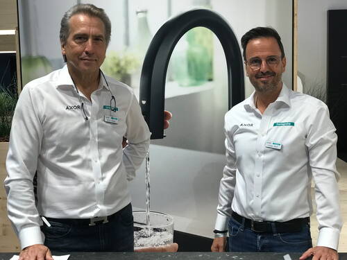 Zwei Mitarbeiter eines Armaturenherstellers stehen in weißer Firmenkleidung vor einem Messestand mit großem Motiv einer laufenden Küchenarmatur.
