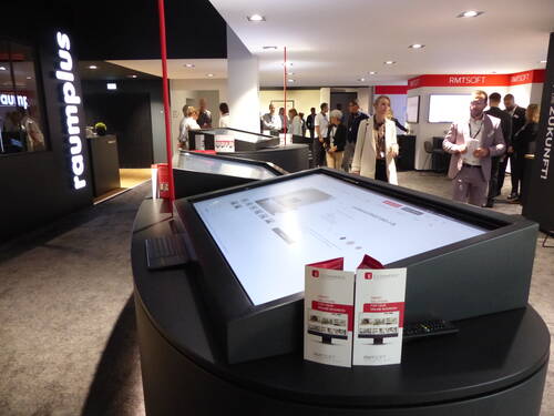 Messebesucher betrachten große interaktive Touchscreens an einem modern gestalteten Messestand mit E‑Commerce‑Flyern im Vordergrund.