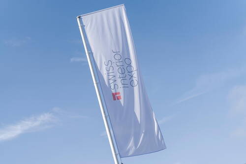 Weiße Fahne mit Aufschrift „Swiss Interior Expo“ weht an hohem Mast vor klarem blauen Himmel