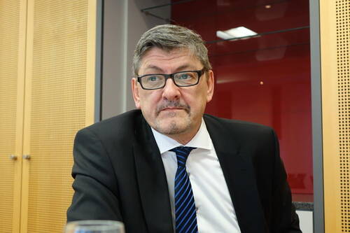 Mittelalterlicher Mann im Anzug mit Brille und nachdenklichem Blick sitzt in einem modernen Konferenzraum mit roten und holzfarbenen Wänden.