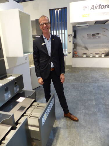 Geschäftsmann im Anzug präsentiert in einer modernen Ausstellungshalle ein offenes CONCEPTO-Küchenmodul mit Schubladensystem.