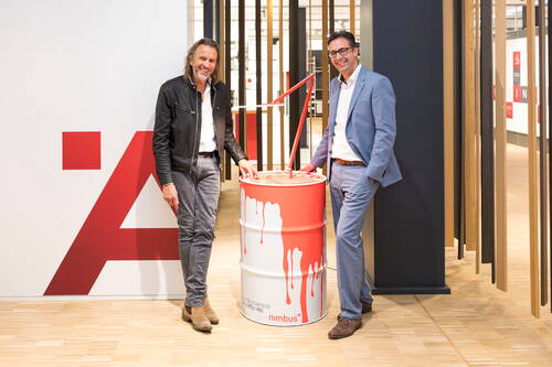 Zwei Männer in Businesskleidung präsentieren in einem modernen Showroom ein rot-weiß lackiertes Ölfass mit integrierter roter Designleuchte.