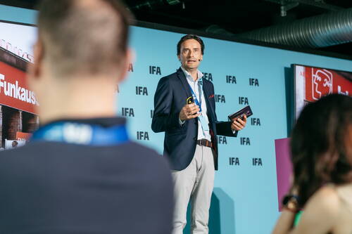 Mann im Anzug hält Präsentation mit Mikrofon und Fernbedienung auf einer IFA‑Bühne vor sitzendem Publikum.