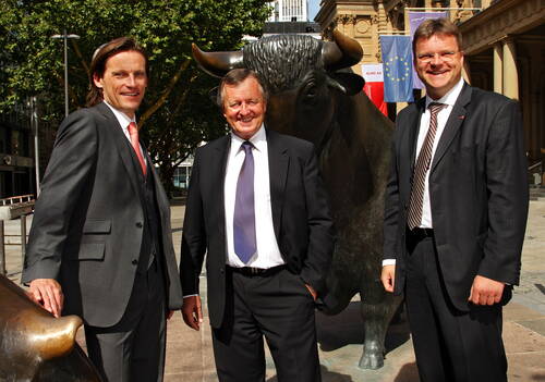 Drei Geschäftsleute in Anzügen posieren lächelnd vor der Bullen-Skulptur an der Frankfurter Börse im Sonnenschein.