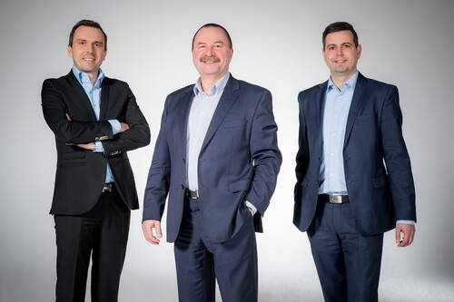 Drei lächelnde Männer in Business-Anzügen posieren vor neutralem Hintergrund als professionelles Teamporträt