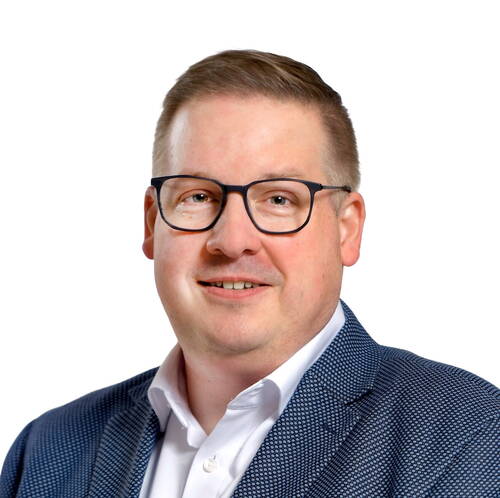 Mittelalterlicher Mann mit Brille und kurzem Haar lächelt in die Kamera, trägt ein weißes Hemd und ein blau gemustertes Sakko vor weißem Hintergrund.