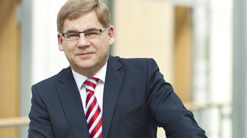 Mittelalterlicher Mann im dunklen Anzug mit rot-weiß gestreifter Krawatte lehnt lächelnd in einem hellen Bürogebäude