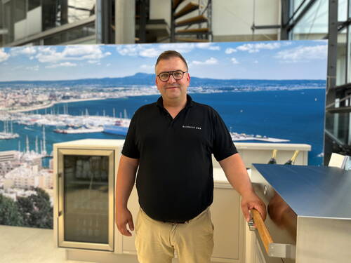 Mann in schwarzem Poloshirt lehnt an einer Edelstahl-Outdoorküche in modernem Ausstellungsraum mit großem Hafen-Panoramabild im Hintergrund