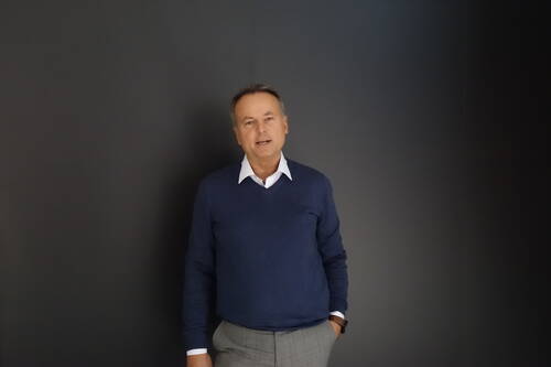 Mittelalterlicher Mann im blauen Pullover und grauer Stoffhose steht entspannt mit einer Hand in der Hosentasche vor einer dunklen, einfarbigen Wand