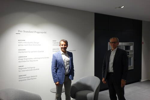 Zwei Geschäftsmänner stehen in einem modernen Showroom vor einer Wand mit dem Schriftzug „Der Standard Poggenpohl“ und Design-Leitlinien, neben grauen Sesseln und einer integrierten Einbauküche.