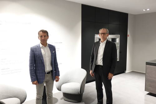 Zwei Männer in Businesskleidung stehen in einem modernen Showroom mit Designer-Sesseln und schwarzer Einbauküche im Hintergrund