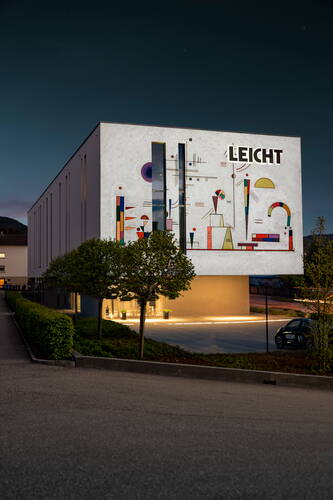 Modernes Firmengebäude mit beleuchtetem LEICHT-Schriftzug und großflächiger abstrakter Wandkunst an der Fassade in der Abenddämmerung