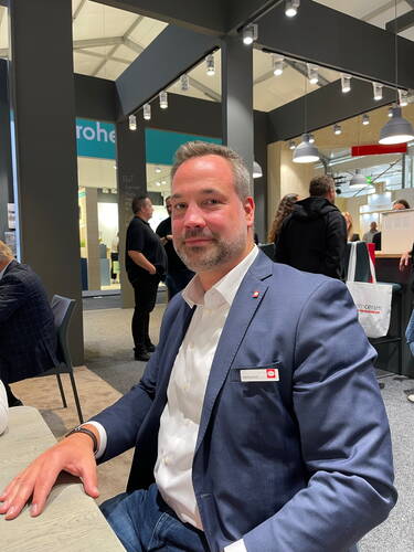 Mann im blauen Anzug sitzt an einem Tisch auf einer Messe und blickt leicht lächelnd in die Kamera, im Hintergrund unterhalten sich mehrere Personen zwischen Messeständen und Deckenstrahlern.