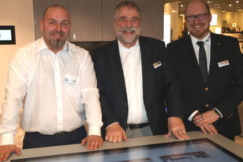 Drei lächelnde Messemitarbeiter stehen nebeneinander hinter einem großen Touchscreen-Tisch in einer modernen Ausstellungshalle.