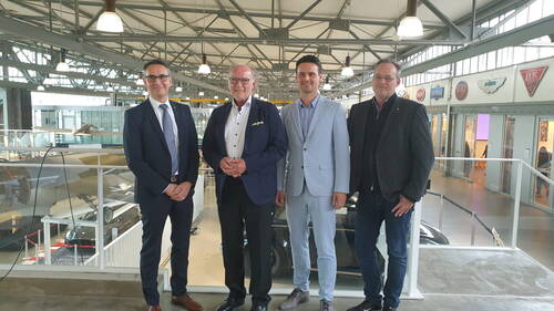 Vier Männer in Anzug und Sakko stehen lächelnd nebeneinander auf einer Galerie in einer hellen Industriehalle mit Oldtimern und Automobilschildern im Hintergrund