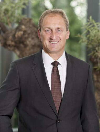 Mittelalterlicher Mann im dunklen Anzug mit Krawatte lächelt freundlich in einer modernen, hellen Umgebung mit großen Fenstern und grünen Pflanzen im Hintergrund