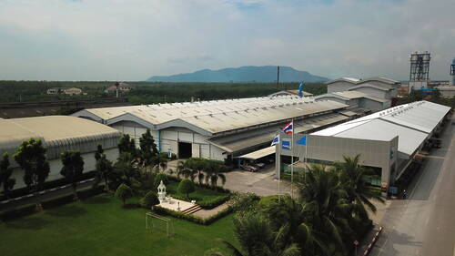 Großes Industrieareal mit langgezogenen Hallen, gepflegtem Garten mit kleinem Fußballfeld und thailändischer Flagge vor grüner Landschaft und entfernten Bergen