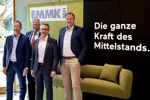 Vier Geschäftsleute in Anzügen posieren vor einem EMMK‑2021‑Banner neben einem schwarzen Hintergrund mit der Aufschrift „Die ganze Kraft des Mittelstands“ und einem grünen Sofa.