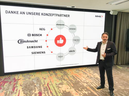 Geschäftsmann im Anzug präsentiert vor großer Leinwand eine Dankesfolie mit Logos der Konzeptpartner AEG, Bosch, Bauknecht, Samsung, Siemens und weiteren Marken bei einem MHK-Group-Seminar.