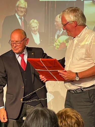 Älterer Mann im Anzug erhält auf einer Bühne von einem lächelnden Mann im Hemd ein rotes, verschnürtes Präsent, während im Hintergrund eine Projektionswand mit einem ähnlichen Bild zu sehen ist.