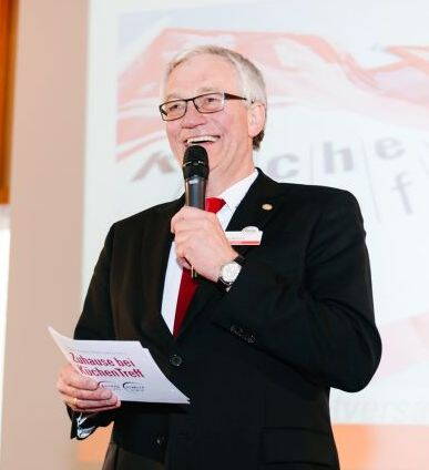 Älterer Mann im Anzug mit roter Krawatte spricht lächelnd in ein Mikrofon und hält Moderationskarten bei einer Veranstaltung auf einer Bühne