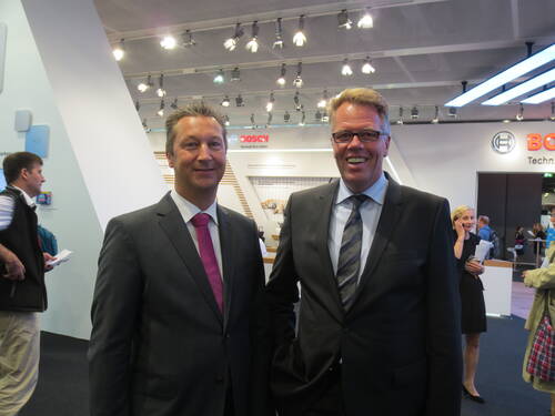 Zwei Geschäftsmänner im Anzug lächeln in einer hell beleuchteten Messehalle mit Bosch-Ausstellungsstand im Hintergrund.