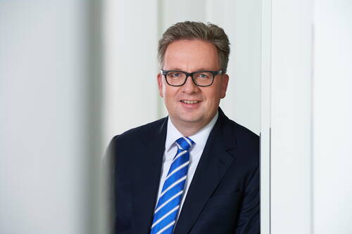 Geschäftsmann mit Brille im dunklen Anzug und blau-weiß gestreifter Krawatte lächelt vor heller, moderner Bürowand in die Kamera
