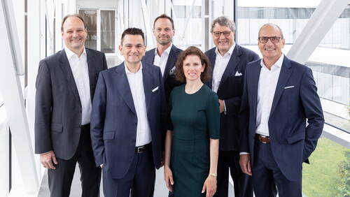 Sechs Business‑Führungskräfte in Anzügen und einem dunkelgrünen Kleid stehen lächelnd in einem hellen verglasten Bürogang und blicken direkt in die Kamera.
