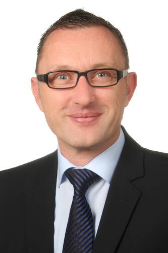 Mittelalterlicher Mann mit Kurzhaarfrisur und Brille lächelt leicht in die Kamera, trägt Anzug mit hellblauem Hemd und dunkelblauer Krawatte vor neutralem, hellem Hintergrund