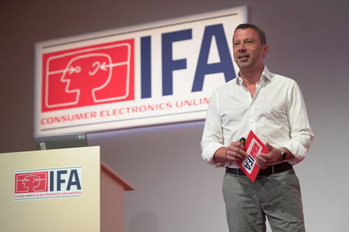 Moderator in weißem Hemd präsentiert auf einer IFA-Bühne vor großem Logo „Consumer Electronics Unlimited“ und hält Mikrofon sowie IFA-Unterlagen in der Hand