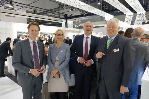 Vier Geschäftsleute in formeller Kleidung lächeln in einer Messehalle in die Kamera, während im Hintergrund zahlreiche Besucher an Stehtischen Gespräche führen.