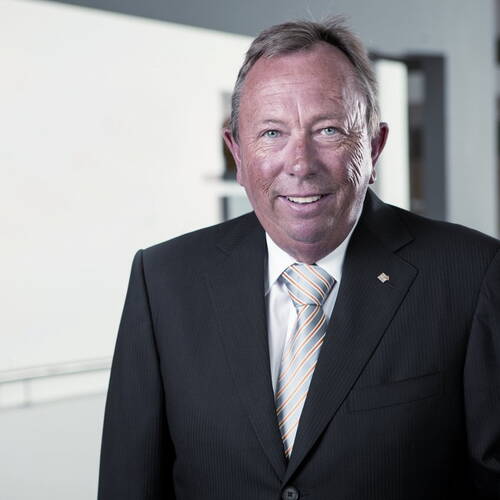 Älterer Mann im dunklen Anzug mit gestreifter Krawatte lächelt in moderner Büro- oder Konferenzumgebung in die Kamera.