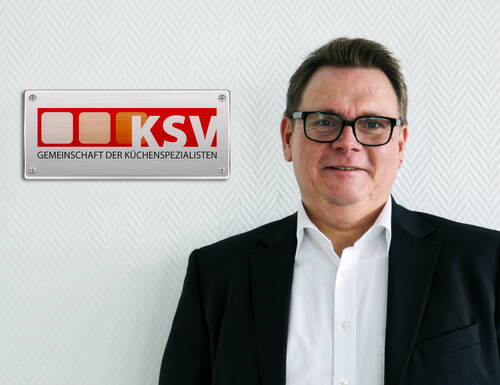 Mann im Anzug lächelt vor heller strukturierten Wand mit KSV-Logo der Gemeinschaft der Küchenspezialisten links neben ihm