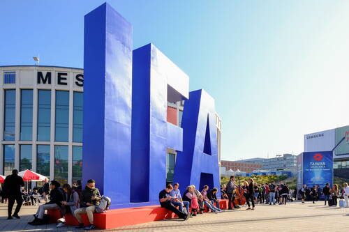 Menschen sitzen und stehen vor einem riesigen blauen IFA-Schriftzug auf dem Messegelände bei sonnigem Wetter
