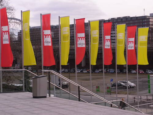 Bunte rot-gelbe Fahnen mit den Logos von „imm cologne“ und „Living Kitchen“ wehen vor einem Messegebäude mit Blick auf Parkplatz und Stadtarchitektur.