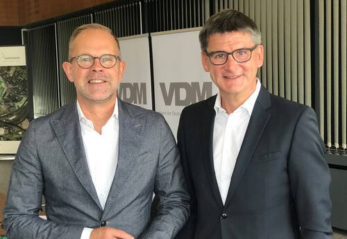 Zwei lächelnde Männer im Anzug stehen nebeneinander vor VDM-Rollups in einem modernen Konferenzraum.