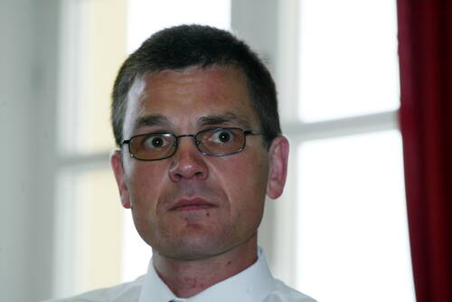 Mittelalterlicher Mann mit Brille und weißem Hemd blickt ernst in die Kamera, im Hintergrund große helle Fenster und ein roter Vorhang.